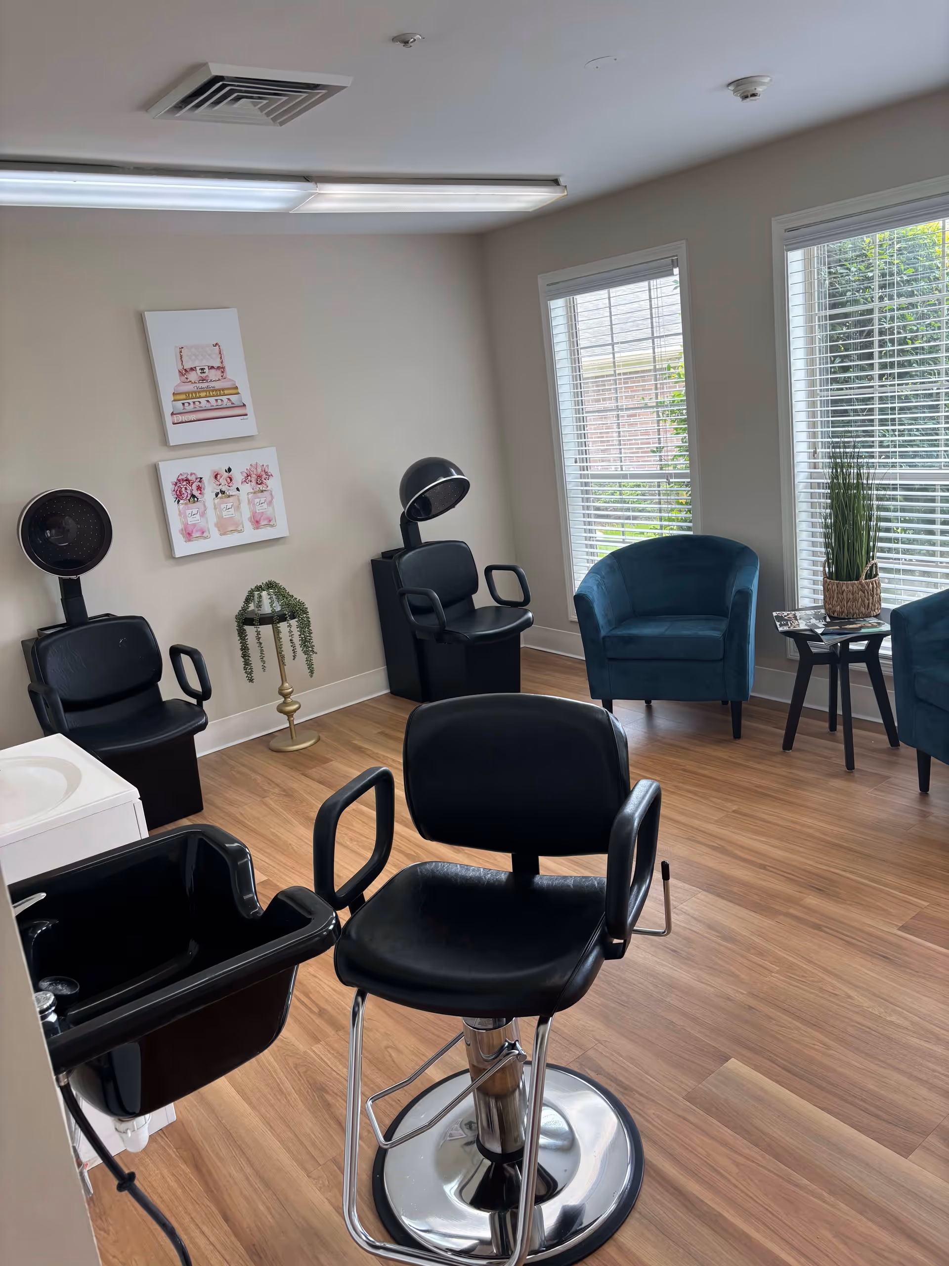 A bright room with wooden flooring featuring a black salon chair with a footrest and attached sink in the foreground. In the background, there are two black hair drying chairs with hooded dryers, two blue upholstered armchairs near large windows with white blinds, a small black side table with a plant and magazines, and two framed pictures on the beige wall.