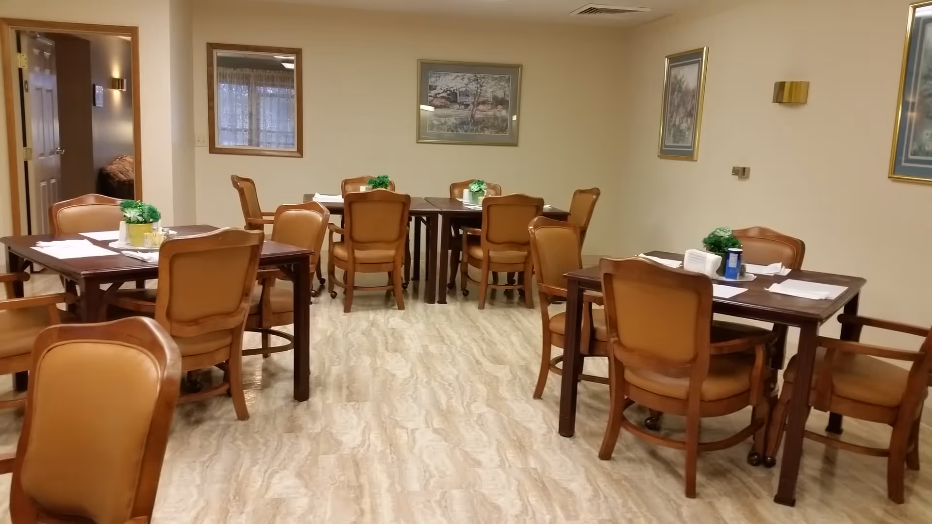 A dining room with several wooden tables and cushioned chairs arranged neatly. Each table has a small green plant centerpiece and place settings with napkins. The room has light-colored walls adorned with framed artwork and a window with curtains. The floor has a light marble pattern.