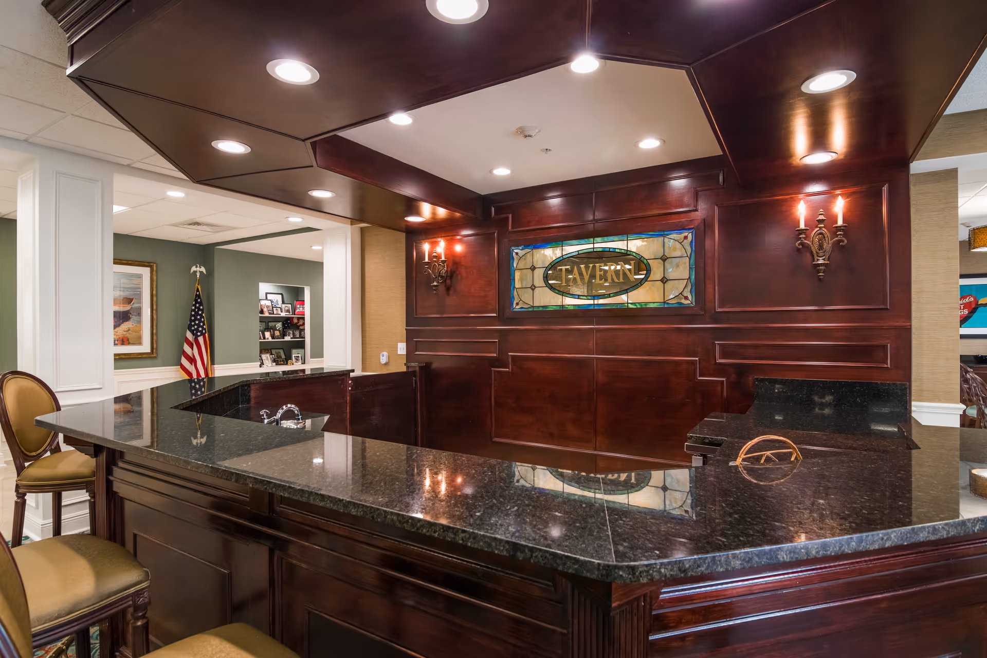 Interior view of a reception or bar area with dark wood paneling and a black granite countertop. The back wall features a stained glass sign that reads 'TAVERN' and two wall sconces with lit candles. There are chairs with green cushions around the counter, and an American flag is visible in the adjacent room.