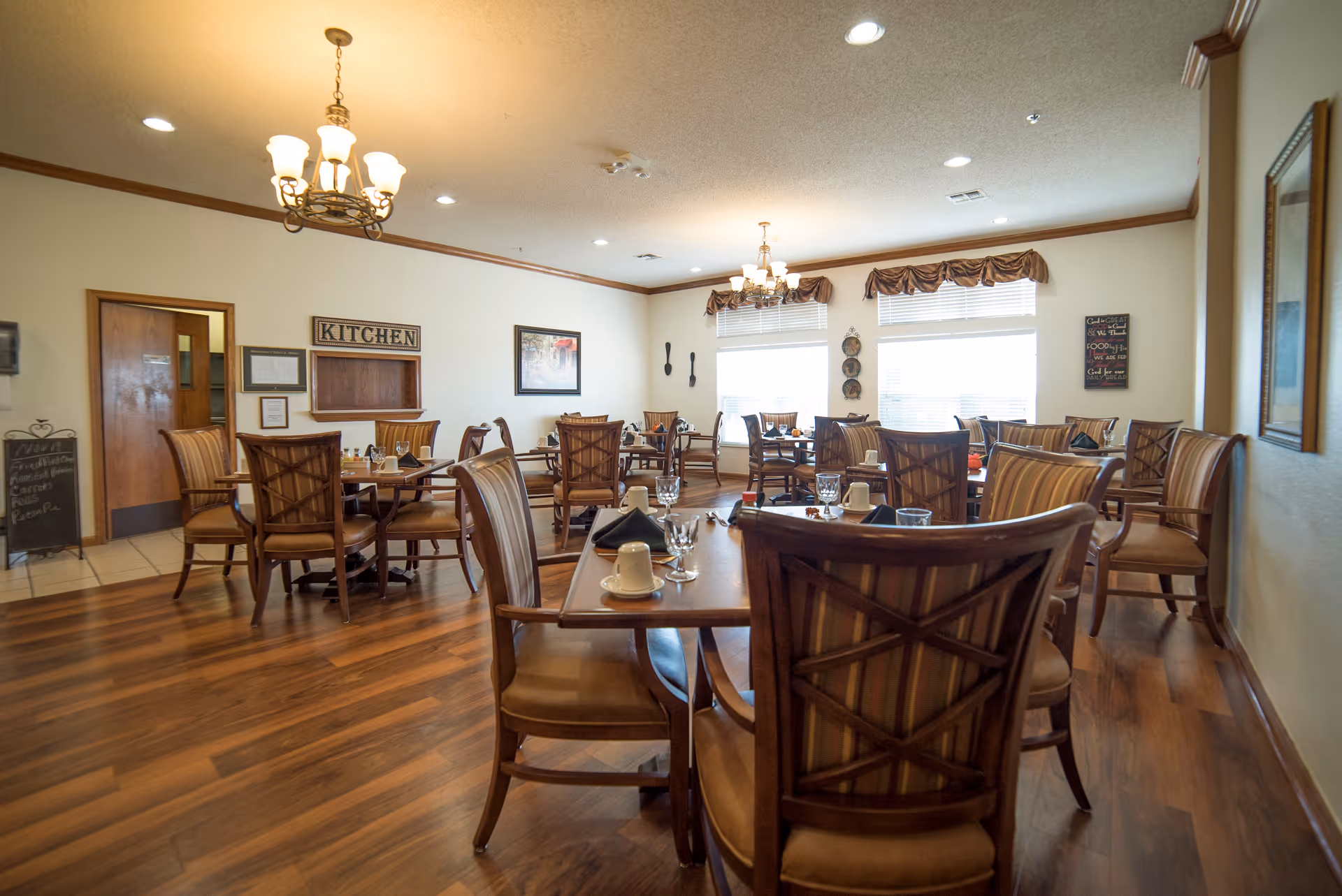 A dining room with multiple wooden tables and chairs arranged neatly. The tables are set with cups, glasses, and napkins. There are two large windows with valances letting in natural light. On the wall, there is a sign that says 'KITCHEN' above a serving window, and decorative items including framed pictures and wall hangings. The floor is wooden, and the ceiling has recessed lighting and chandeliers.