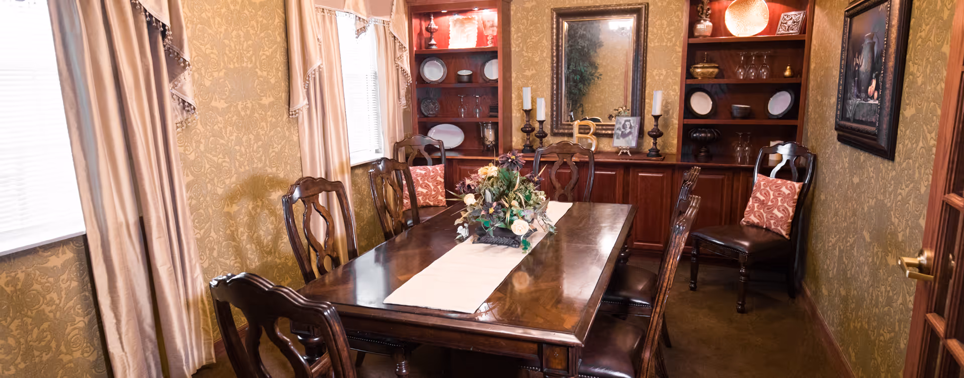 A traditional dining room with a polished wooden table and six matching chairs, a floral centerpiece on a table runner, beige patterned wallpaper, two windows with beige curtains, built-in wooden shelves displaying plates and decorative items, a mirror, candles, and framed artwork on the walls.