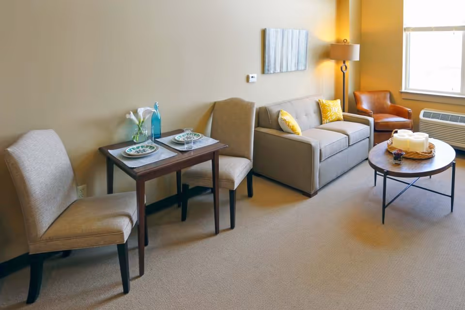 A cozy living room area with a small dining table set for two, a beige sofa with yellow patterned pillows, a brown leather armchair, a round coffee table with candles and a small decorative item, a floor lamp, and a window with an air conditioning unit below it.
