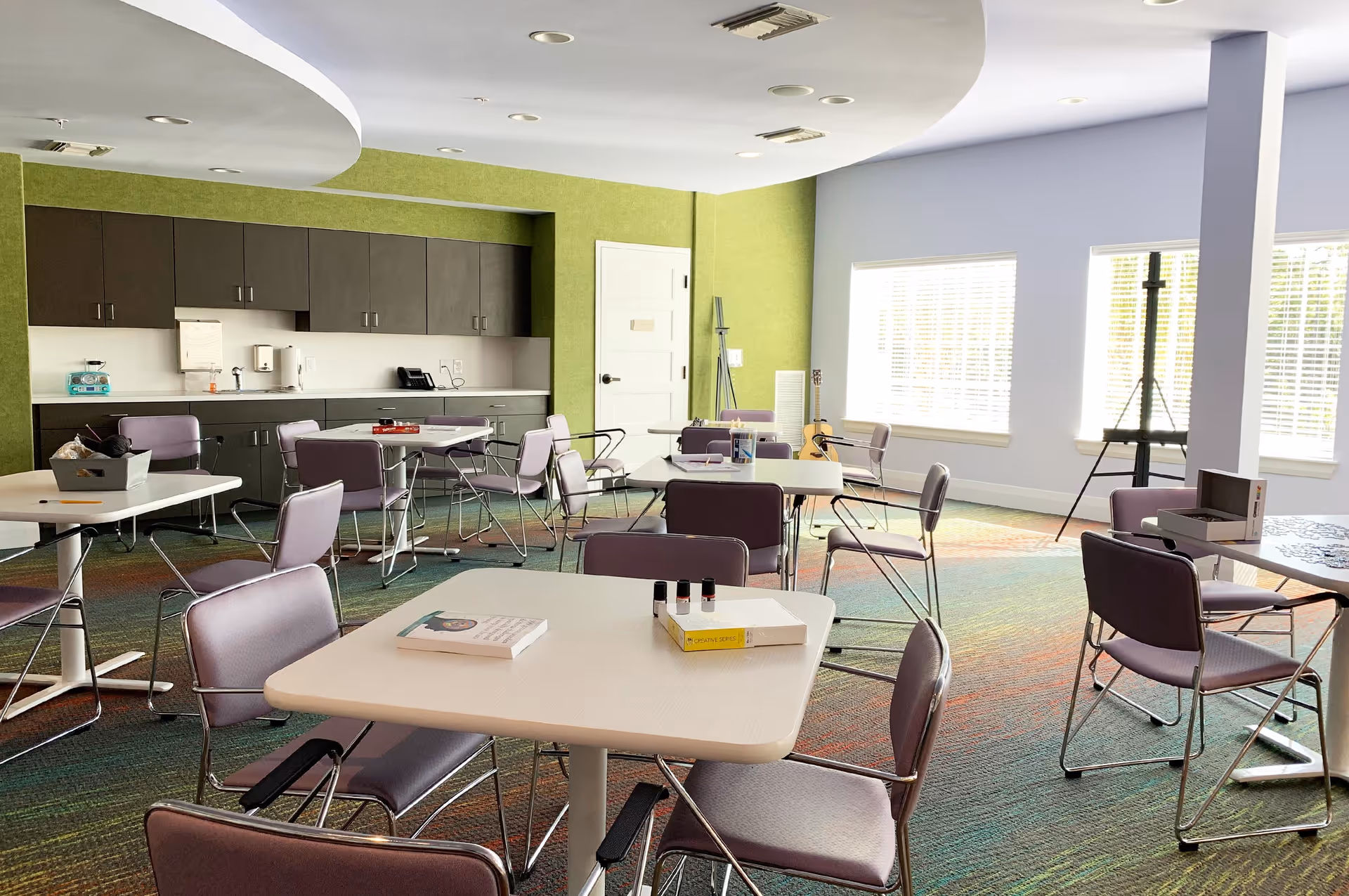 Bright multipurpose dining/activity room with several square tables and purple chairs, a kitchenette against a green wall, and large windows.