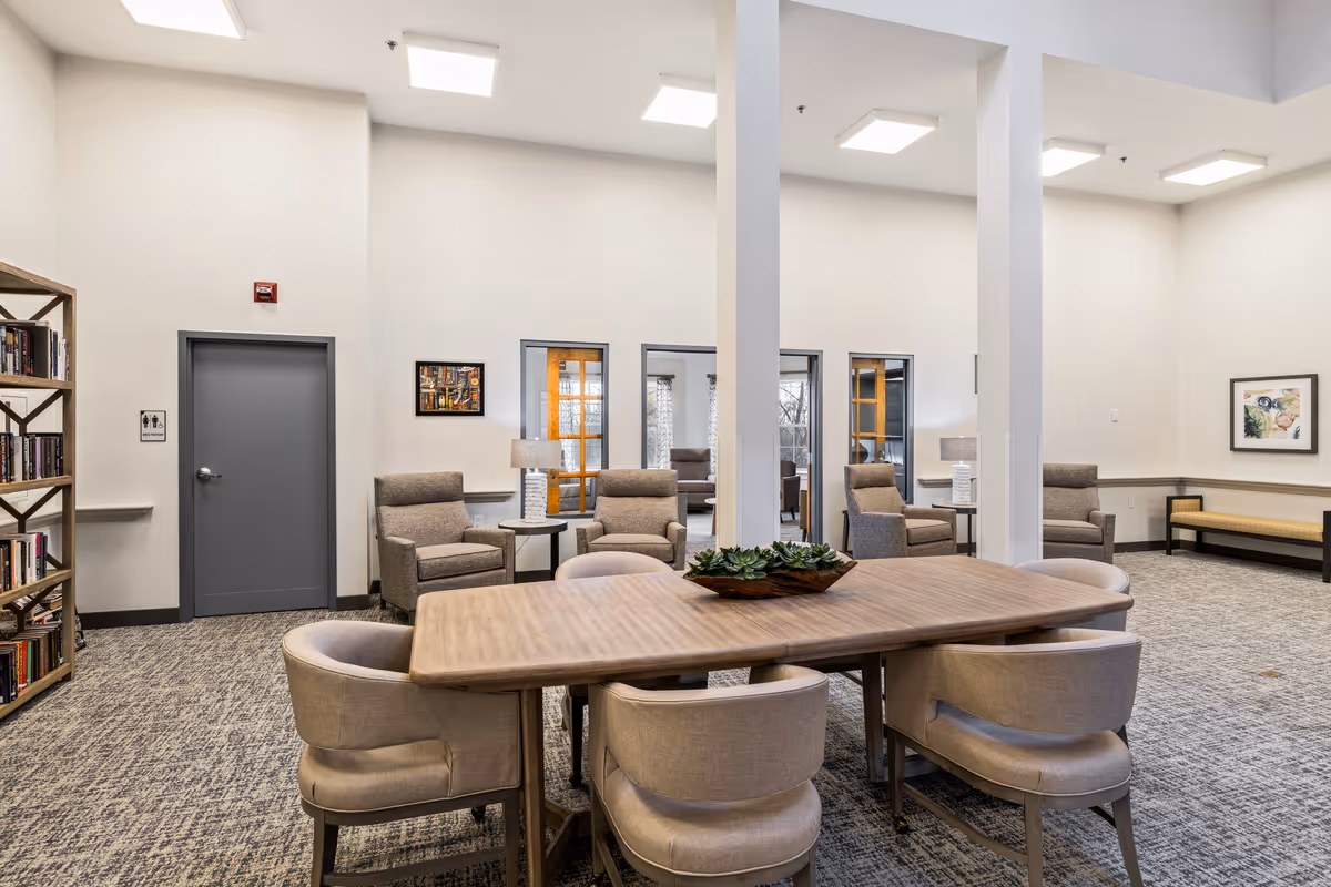 Bright communal lounge with a wooden table surrounded by upholstered chairs, armchairs, and bookshelves in a senior living facility.