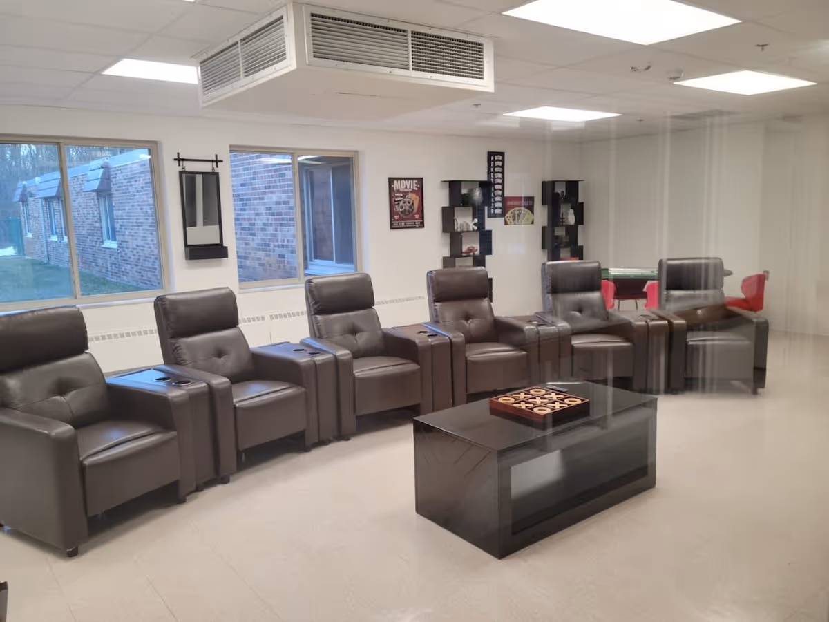 A lounge area with six brown leather recliner chairs arranged in a row facing a black coffee table with a tic-tac-toe game on top. The room has white walls, large windows showing a brick exterior, and shelves with decorative items and posters on the wall. The ceiling has recessed lighting and an air vent.