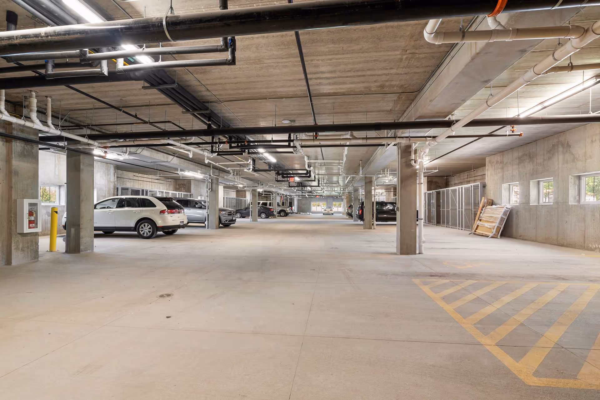 Indoor parking garage with several parked cars along the left side and concrete pillars supporting the ceiling. The ceiling has exposed pipes and fluorescent lighting. There are small windows on the right wall and a fire extinguisher mounted on a pillar on the left.
