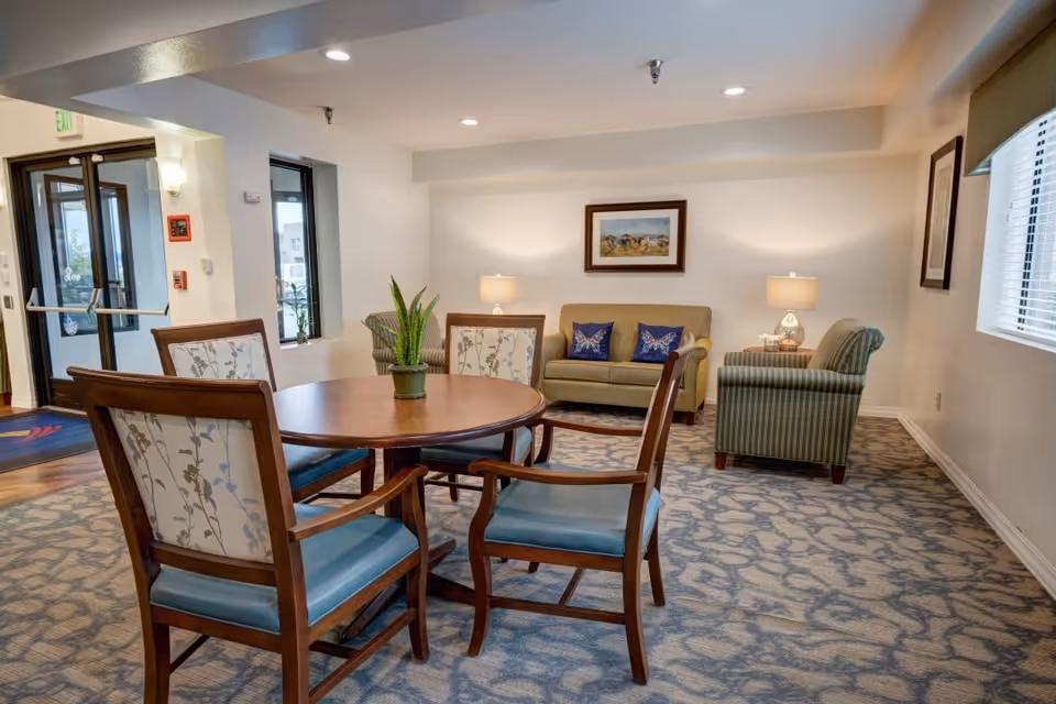 A cozy common area in a senior living facility featuring a round wooden table with four cushioned chairs, a small potted plant centerpiece, a beige loveseat with two butterfly-themed pillows, a striped armchair, two table lamps, framed artwork on the walls, and large windows letting in natural light.