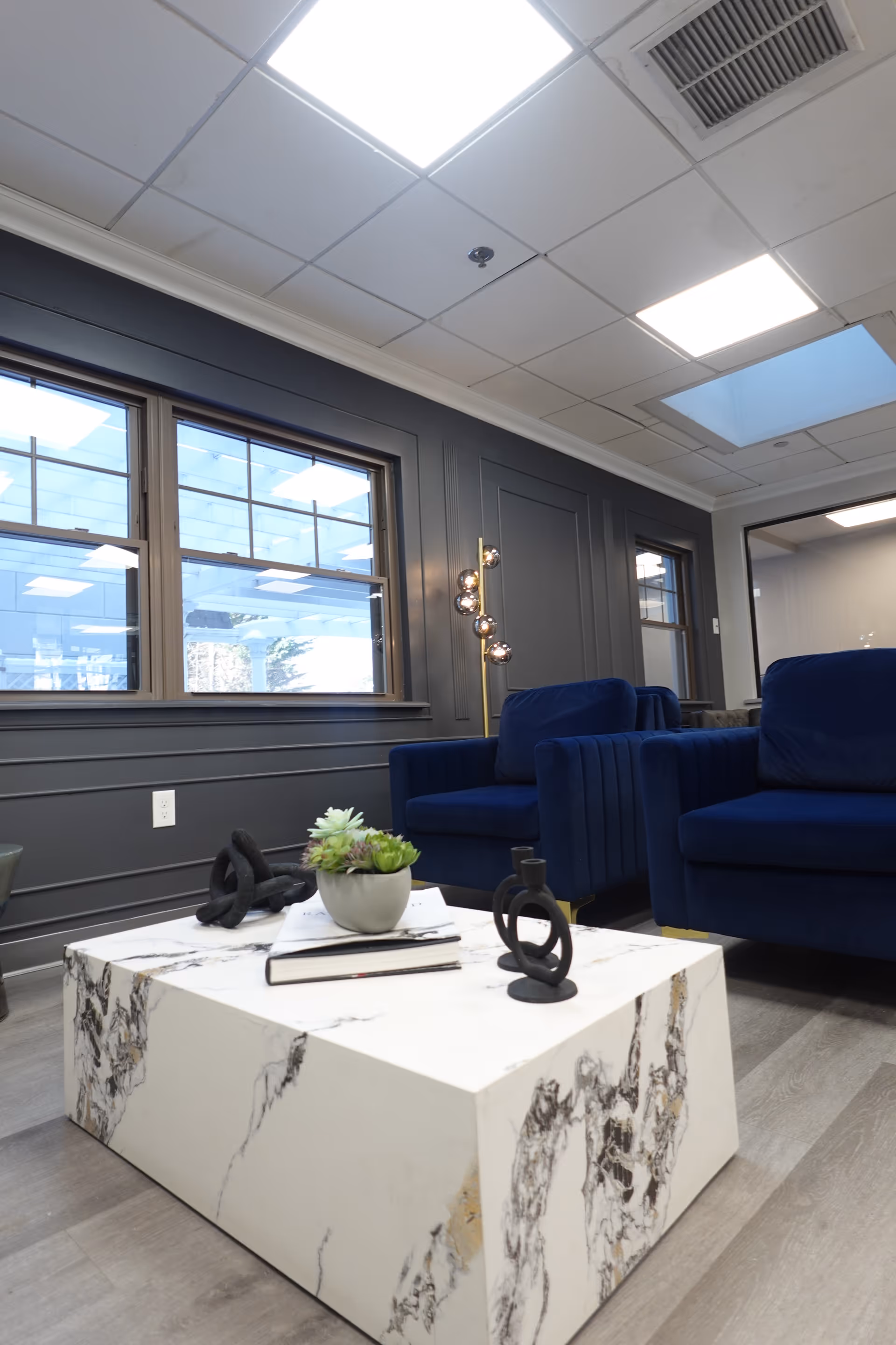 Modern interior lounge area with dark blue armchairs, a white marble-patterned coffee table adorned with a small potted succulent, decorative objects, and a book. The room features dark gray paneled walls, large windows, and a ceiling with recessed lighting and a skylight.