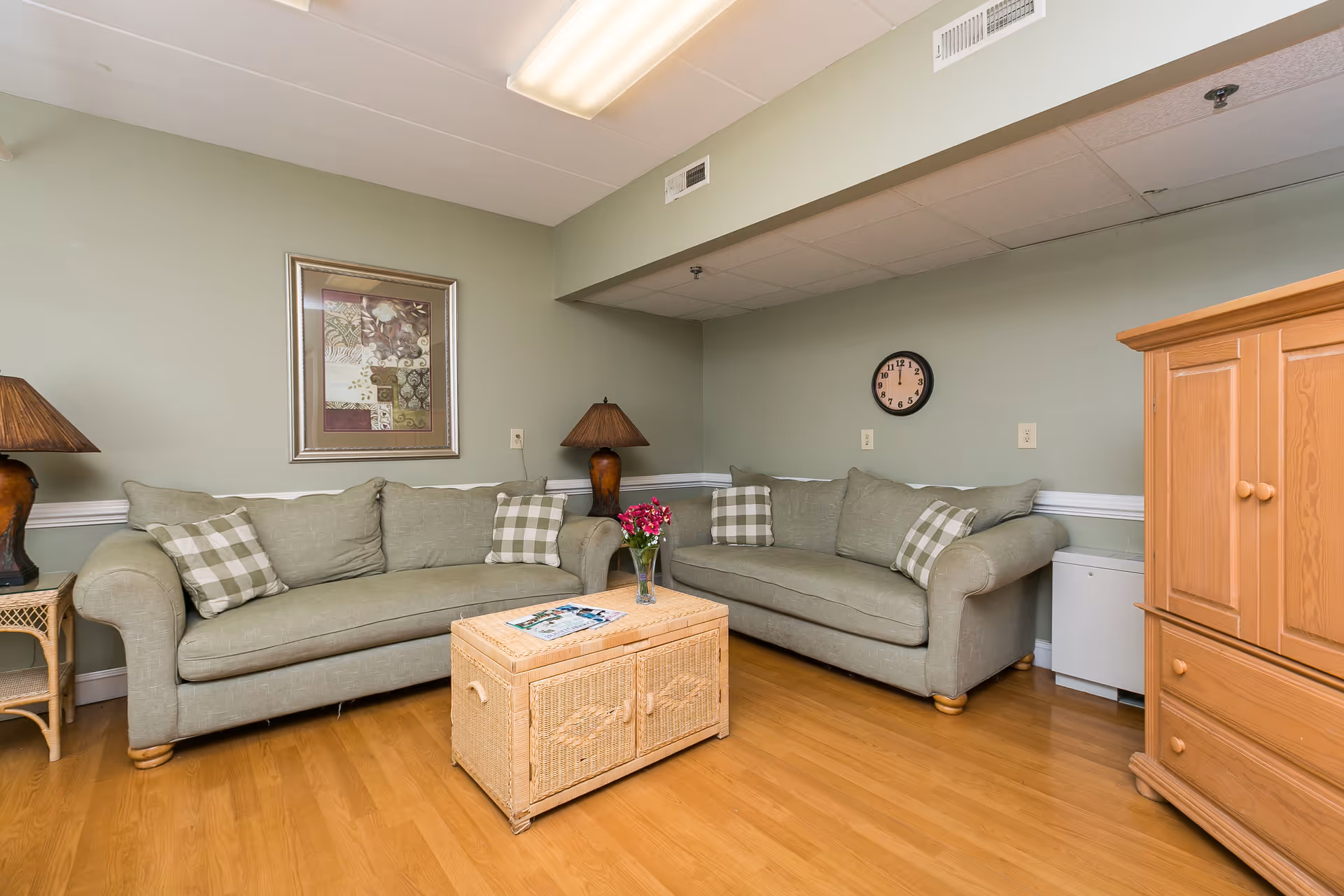 A cozy living room with two light green sofas adorned with plaid pillows, a wicker coffee table with a vase of flowers and magazines, two table lamps with brown bases and shades, a framed artwork on the wall, a round clock, and a wooden cabinet on a hardwood floor.