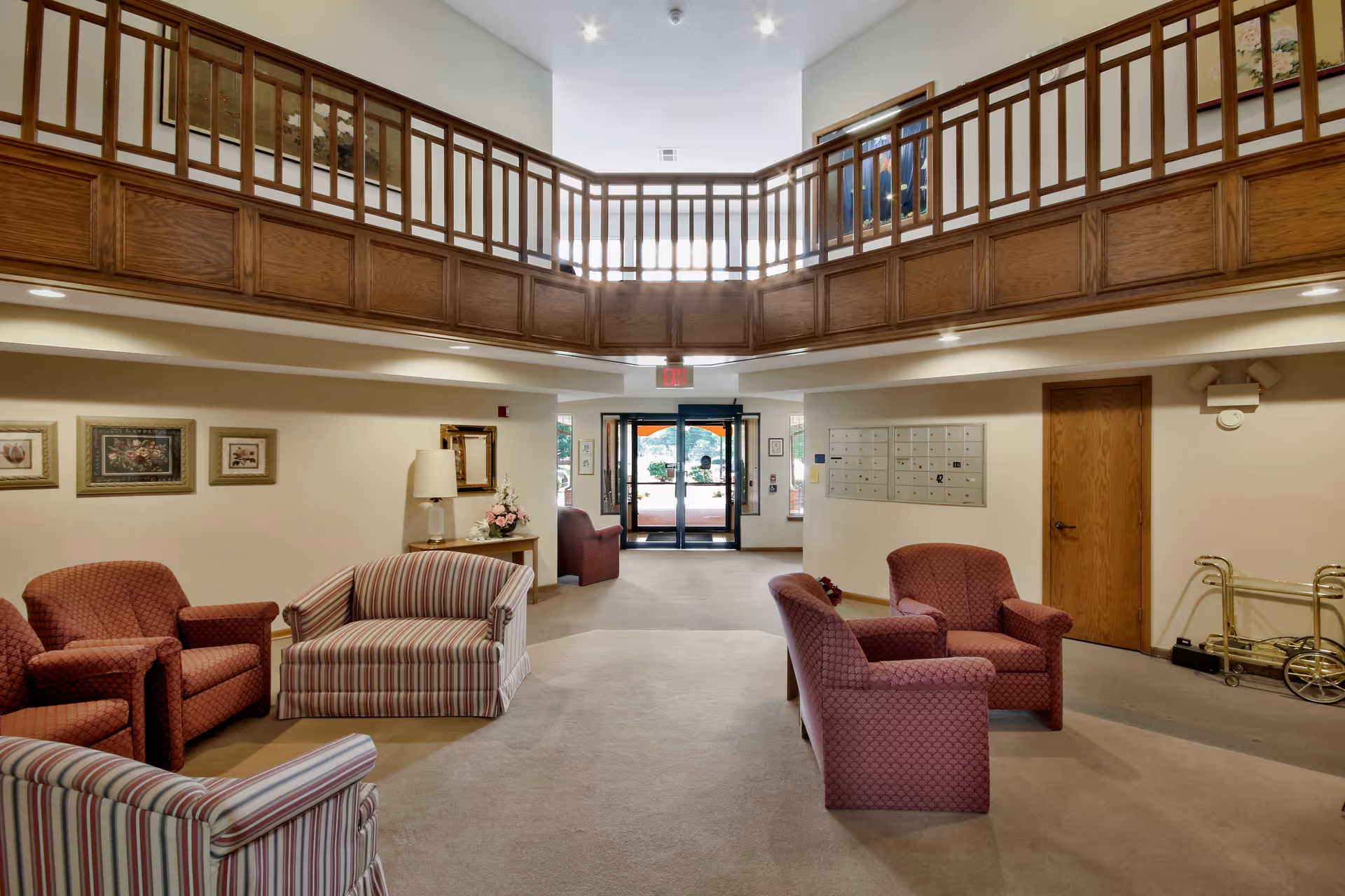 Interior view of a senior living facility lobby area with comfortable upholstered chairs and sofas arranged in seating groups. The space features a high ceiling with a wooden balcony railing above, framed artwork on the walls, a table with a lamp and flower arrangement, and a set of mailboxes on one wall. Double glass doors lead outside, allowing natural light to enter the room.