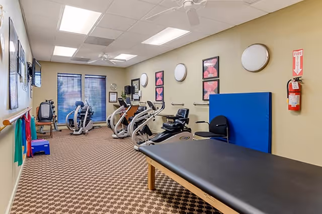 A fitness room in a senior living facility with exercise equipment including stationary bikes and a recumbent bike. The room has beige walls with framed artwork, a patterned carpet, a large window with blinds, and a black padded exercise table in the foreground. There is a fire extinguisher mounted on the wall and ceiling fans overhead.