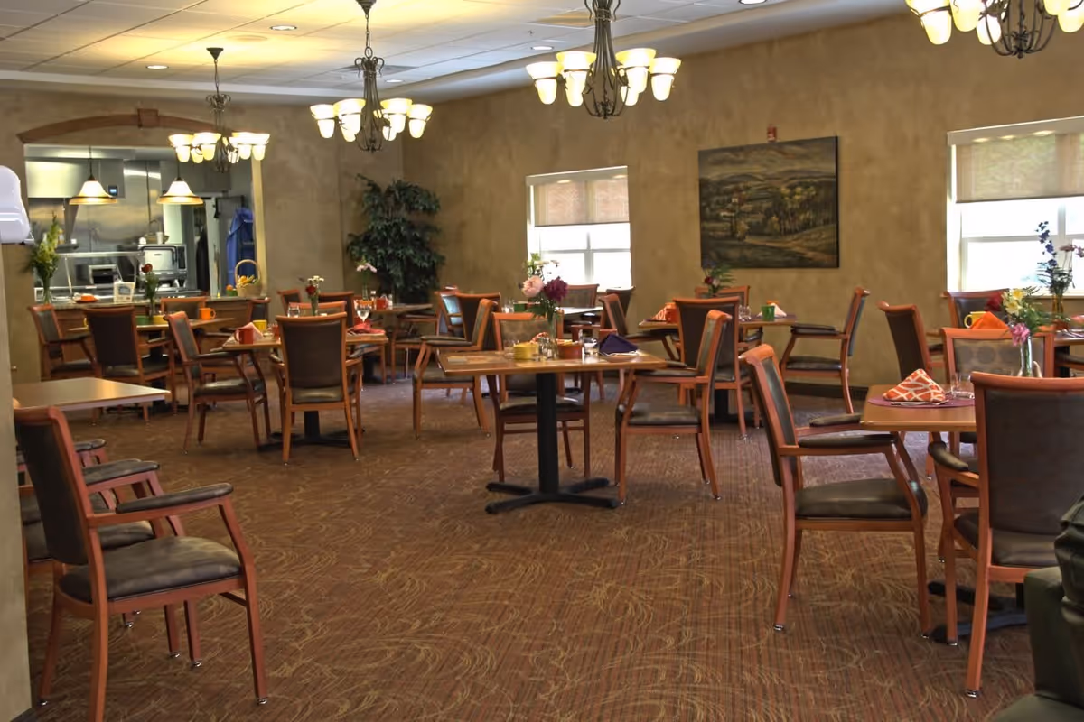 A dining room in a senior living facility with multiple wooden tables and chairs arranged neatly. Each table has a small vase with flowers and folded napkins. The room has warm lighting from chandeliers and windows with blinds. A painting hangs on the wall, and a large plant is placed in the corner near the window. The kitchen area is visible through an open archway.