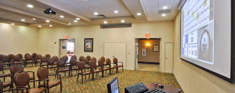 A large room set up with rows of brown chairs facing a projection screen displaying a bingo game. The room has patterned carpet, beige walls, and ceiling lights. There is a podium with a computer and control panel in the foreground.