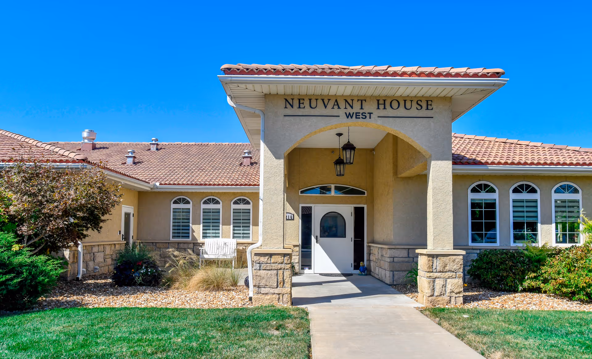 Front exterior view of Neuvant House West building with a covered entrance, beige walls, stone accents, and a red tile roof under a clear blue sky. There are bushes and a green lawn in front of the building.