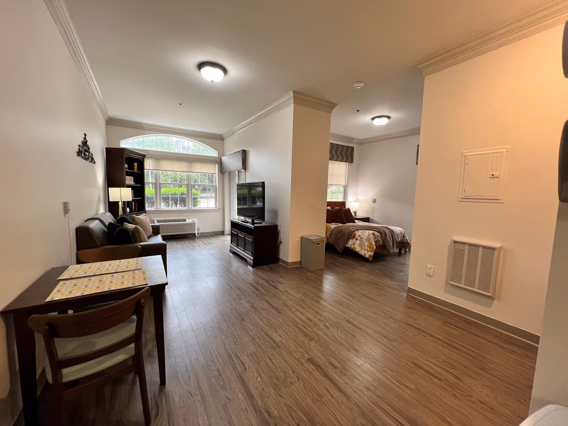 A spacious assisted living apartment featuring a living area with a sofa, wooden bookshelf, and a TV on a stand. There is a dining table with two chairs in the foreground. The bedroom area is partially visible, showing a bed with floral bedding and a nightstand with a lamp. Large windows allow natural light to fill the room, and the flooring is wood-style laminate.
