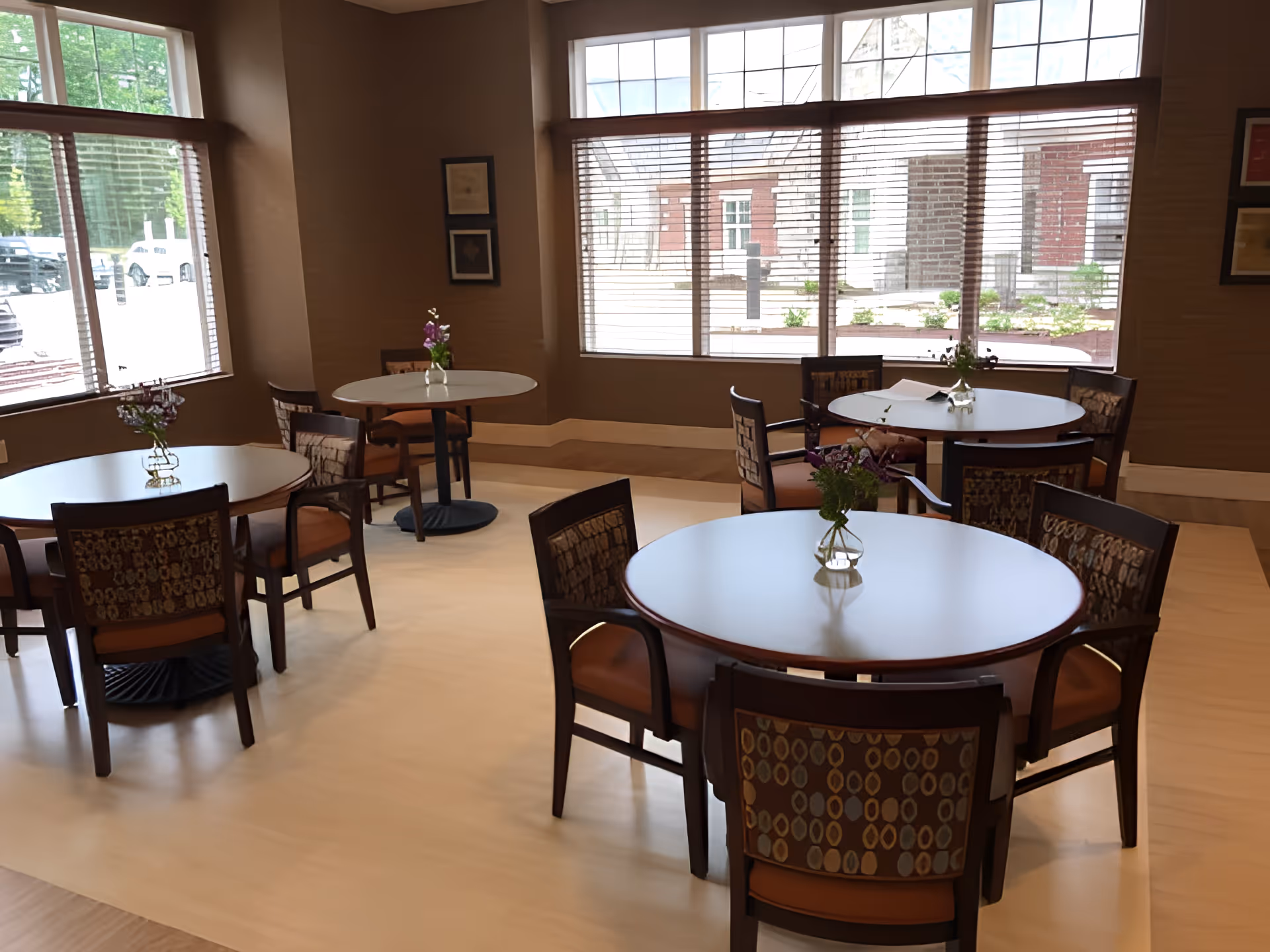 Dining area with round tables and chairs, small vases of flowers, and large windows.
