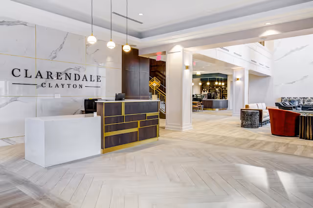 Bright and modern reception area of Clarendale Clayton featuring a white marble wall with the facility name, a sleek reception desk with dark wood and gold accents, pendant lights hanging from the ceiling, and an open seating area with chairs and tables in the background.
