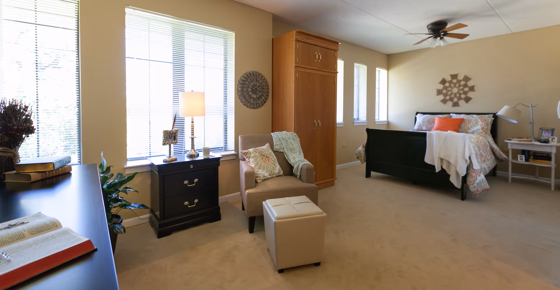 A spacious bedroom with a large bed adorned with patterned bedding and orange accent pillows. There is a beige armchair with a decorative pillow and a throw blanket, accompanied by a small ottoman. The room features multiple windows with blinds, a ceiling fan, a wooden wardrobe, a black nightstand with a lamp, and a side table with a lamp and framed photo. The walls are painted light beige and the floor is carpeted.