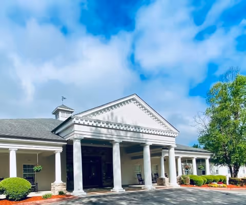 Front exterior view of a building with white columns and a triangular pediment under a partly cloudy blue sky. The entrance is shaded by a covered porch with rocking chairs and hanging plants. There are neatly trimmed bushes and a tree on the right side.