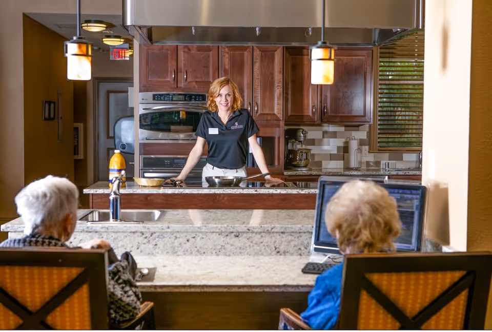 A woman stands behind a kitchen counter smiling, with two elderly women seated on the opposite side. The kitchen features wooden cabinets, a built-in oven, a mixer, and pendant lights hanging from the ceiling. One elderly woman is using a laptop.