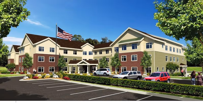 Exterior view of a three-story senior living facility building with a combination of beige and brick facade, surrounded by trees and landscaping. There is a parking lot with several cars in front, an American flag on a flagpole near the entrance, and a few people walking near the building.