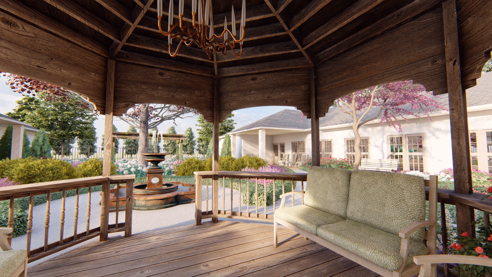 View from inside a wooden gazebo with a cushioned bench, overlooking a landscaped garden with a central water fountain, trees, and flowering plants. In the background, there is a single-story building with large windows and a covered porch area with seating.