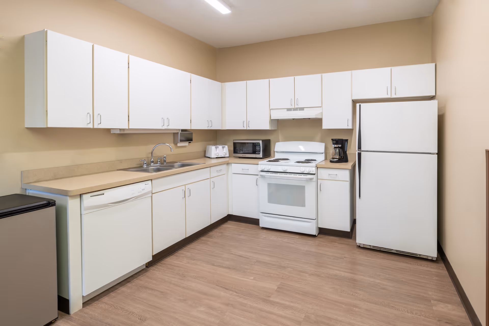 A clean and well-lit kitchen with white cabinets, beige countertops, and light wood flooring. The kitchen features a white stove with an oven, a white refrigerator, a dishwasher, a microwave, a toaster, and a coffee maker. The walls are painted beige.