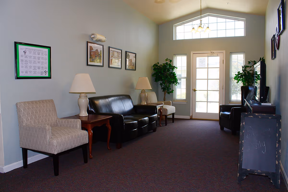 A quiet sitting area with a black leather sofa, two beige armchairs, two table lamps on wooden side tables, framed pictures on the wall, two potted plants near a glass door with windows, and a TV on a stand with a small chalkboard sign beside it.