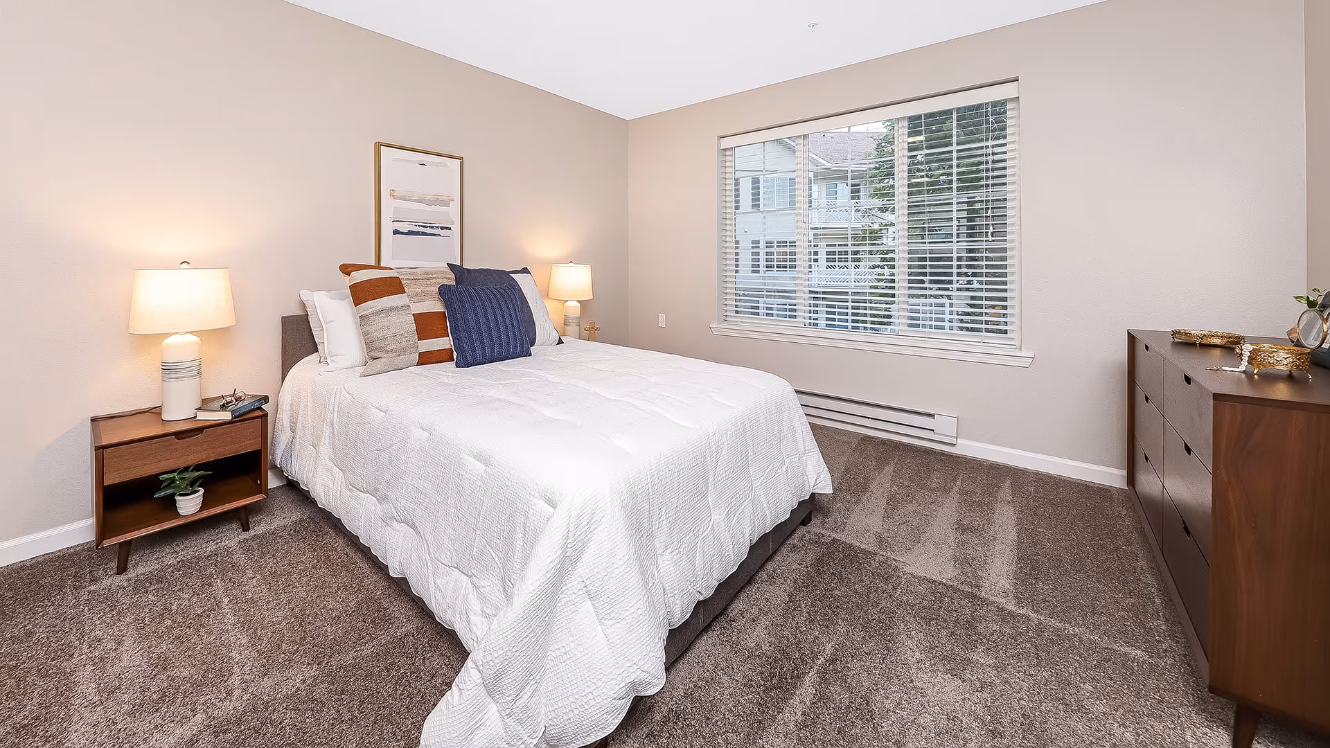 Bright, neatly staged bedroom with a bed, nightstands and lamps, a dresser, and a window with blinds.