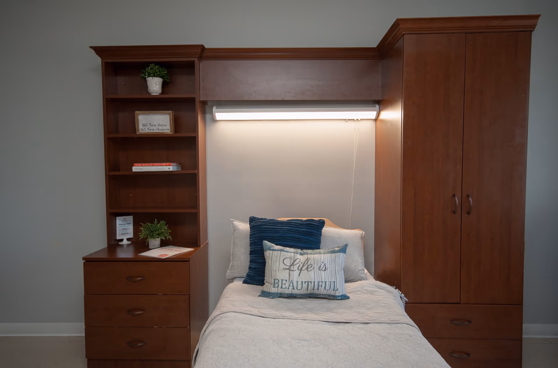 Single bed with decorative pillows including a 'Life is Beautiful' cushion set within wooden built-in shelving and cabinetry under a wall light.