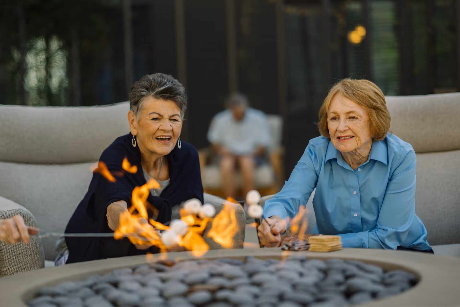 Two elderly women sitting on cushioned outdoor chairs around a fire pit, roasting marshmallows. One woman is wearing a dark shawl and the other is in a light blue shirt. There is a blurred figure in the background also seated.