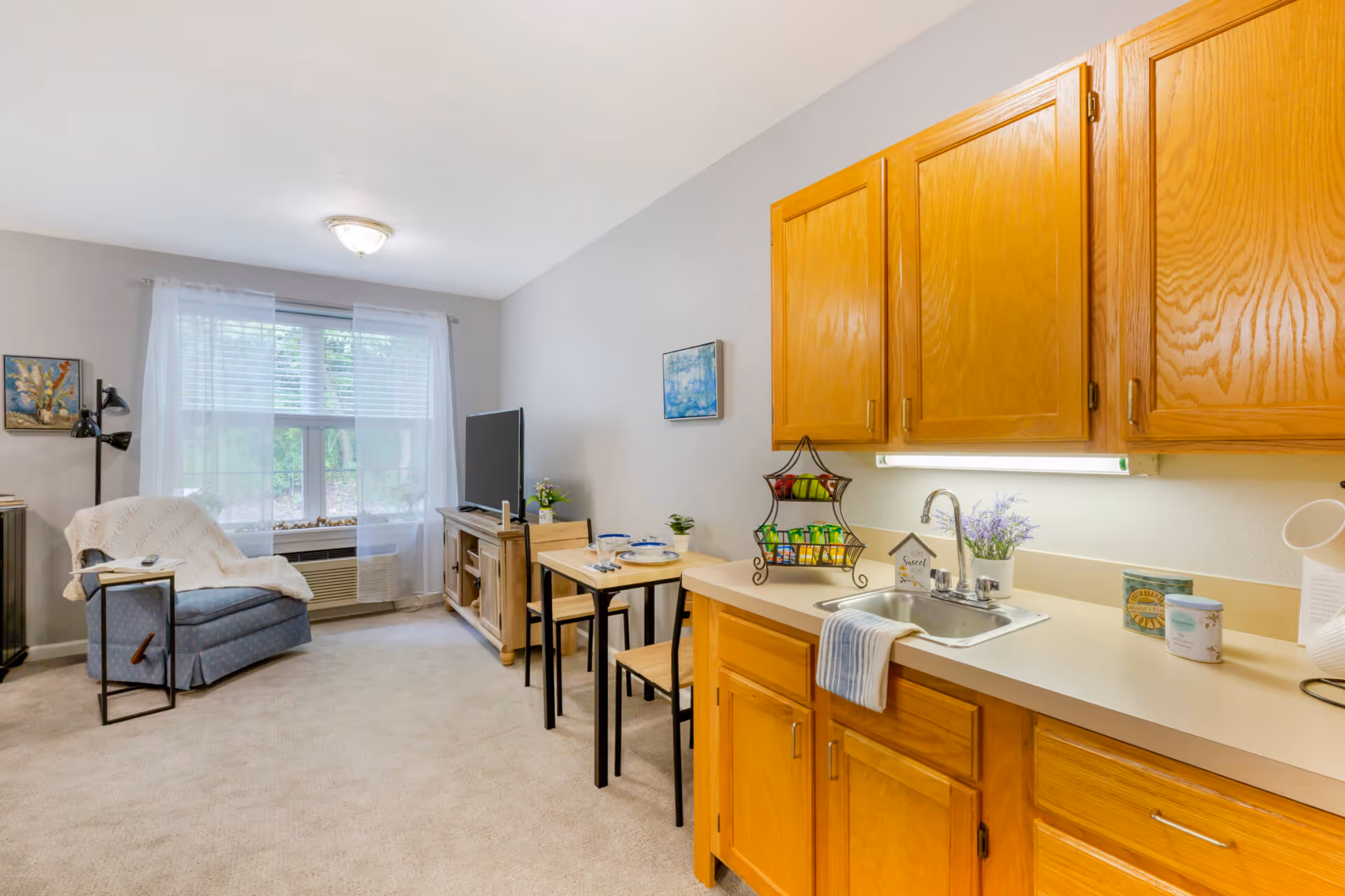A cozy living space featuring a small kitchen area with wooden cabinets, a sink, and countertop. Adjacent to the kitchen is a small dining table set for two. The living area includes a comfortable armchair with a white throw blanket, a floor lamp, a TV on a wooden stand, and a large window with sheer white curtains allowing natural light to fill the room.
