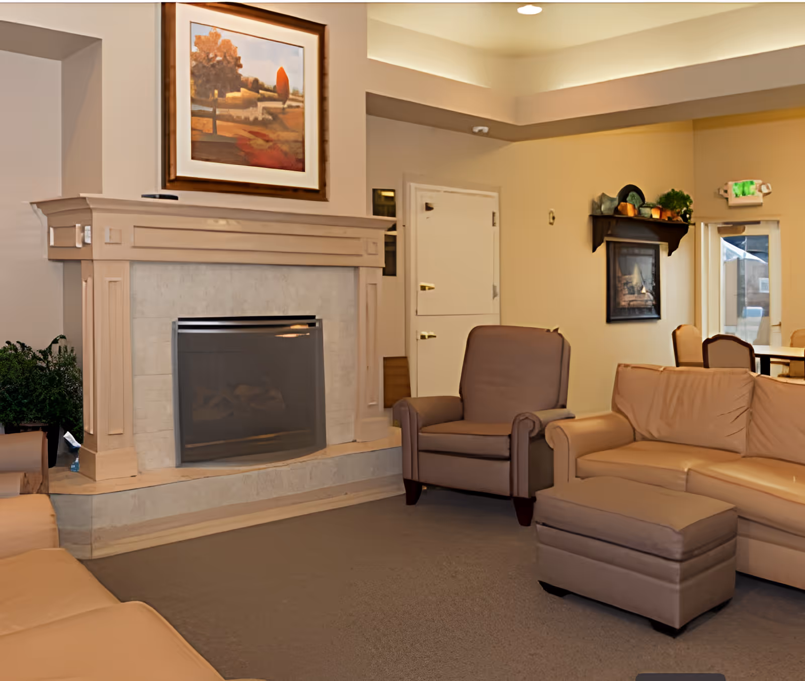 A cozy living room area featuring a beige leather sofa, matching ottoman, and a brown armchair arranged around a fireplace with a light-colored mantel. Above the fireplace is a framed landscape painting. The room has beige walls, a carpeted floor, and a shelf with decorative items on the wall near a door and window.