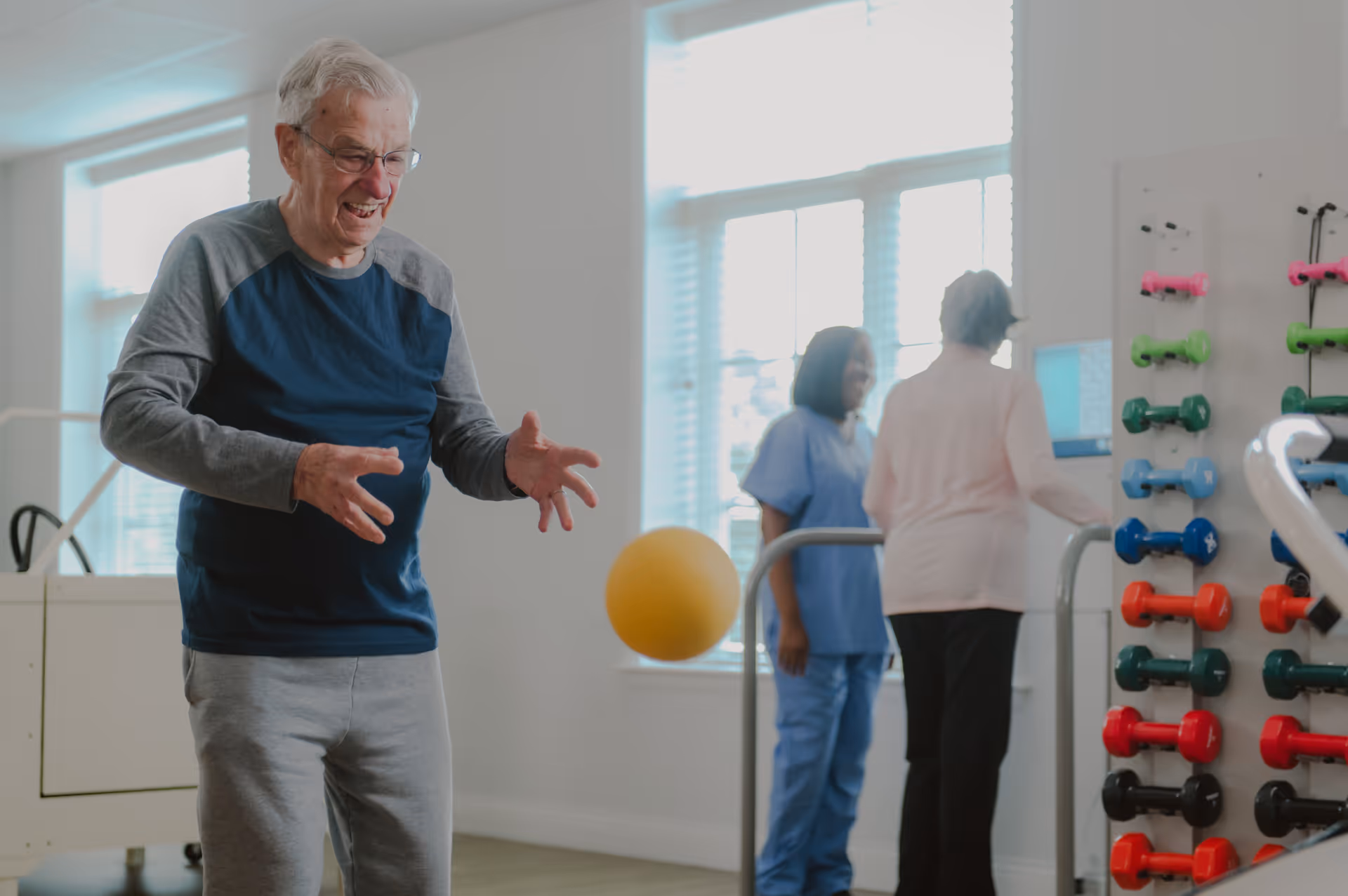 An older man smiles while tossing a yellow exercise ball in a bright rehabilitation room with staff and a rack of colorful dumbbells.