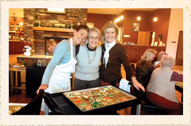 Three smiling women standing behind a cart filled with decorated holiday cookies in a cozy room with a stone fireplace and wooden furniture. In the background, other people are seated at tables engaged in activities.