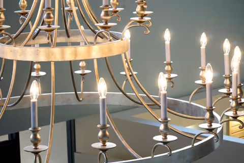 Close-up view of a large decorative chandelier with multiple lit candle-shaped light bulbs and a metallic frame, hanging indoors with a muted green wall in the background.