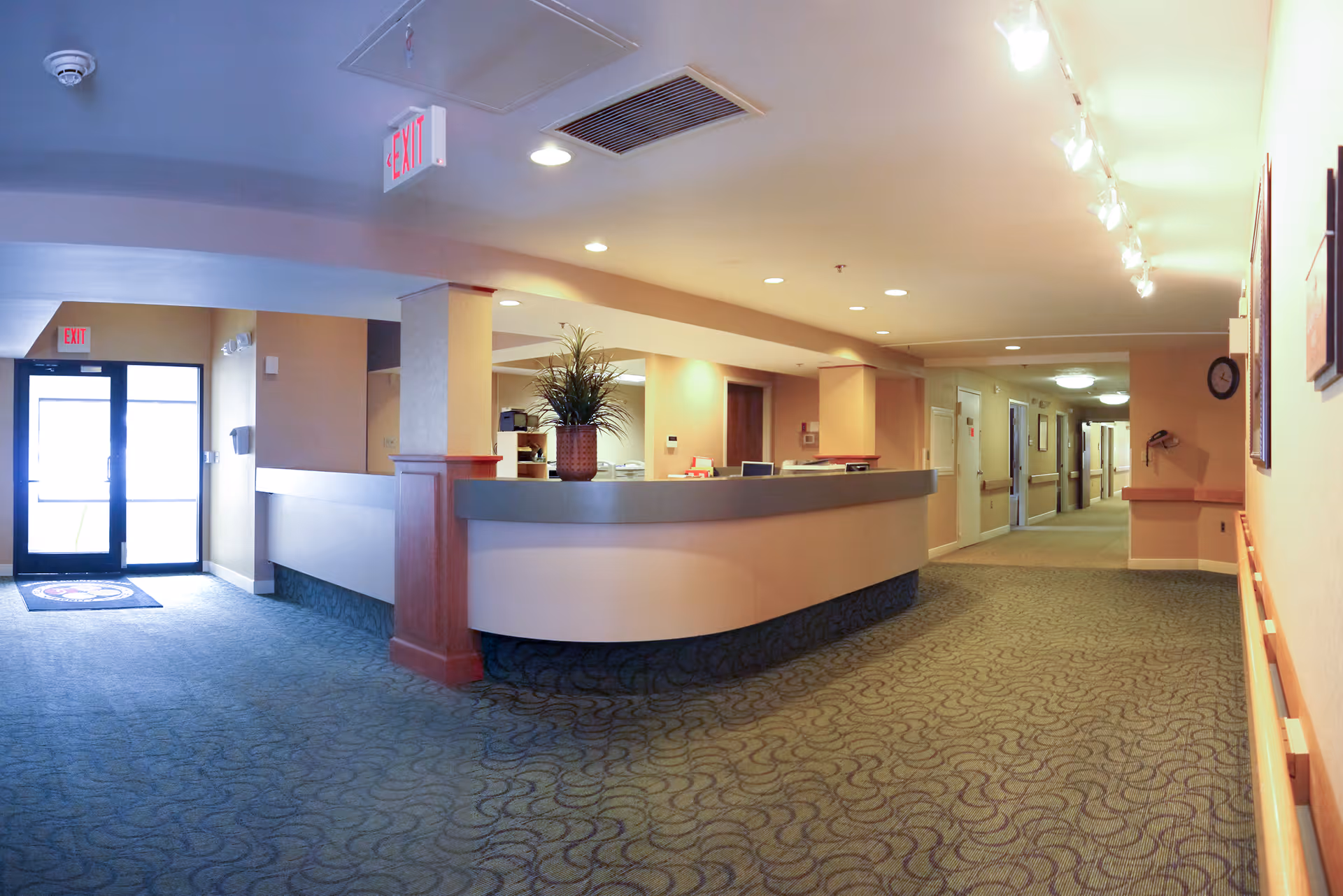 Interior view of a senior living facility hallway with a reception desk on the left side. The hallway is carpeted with patterned flooring and has beige walls with handrails. There are ceiling lights and exit signs visible, along with a potted plant on the reception desk.