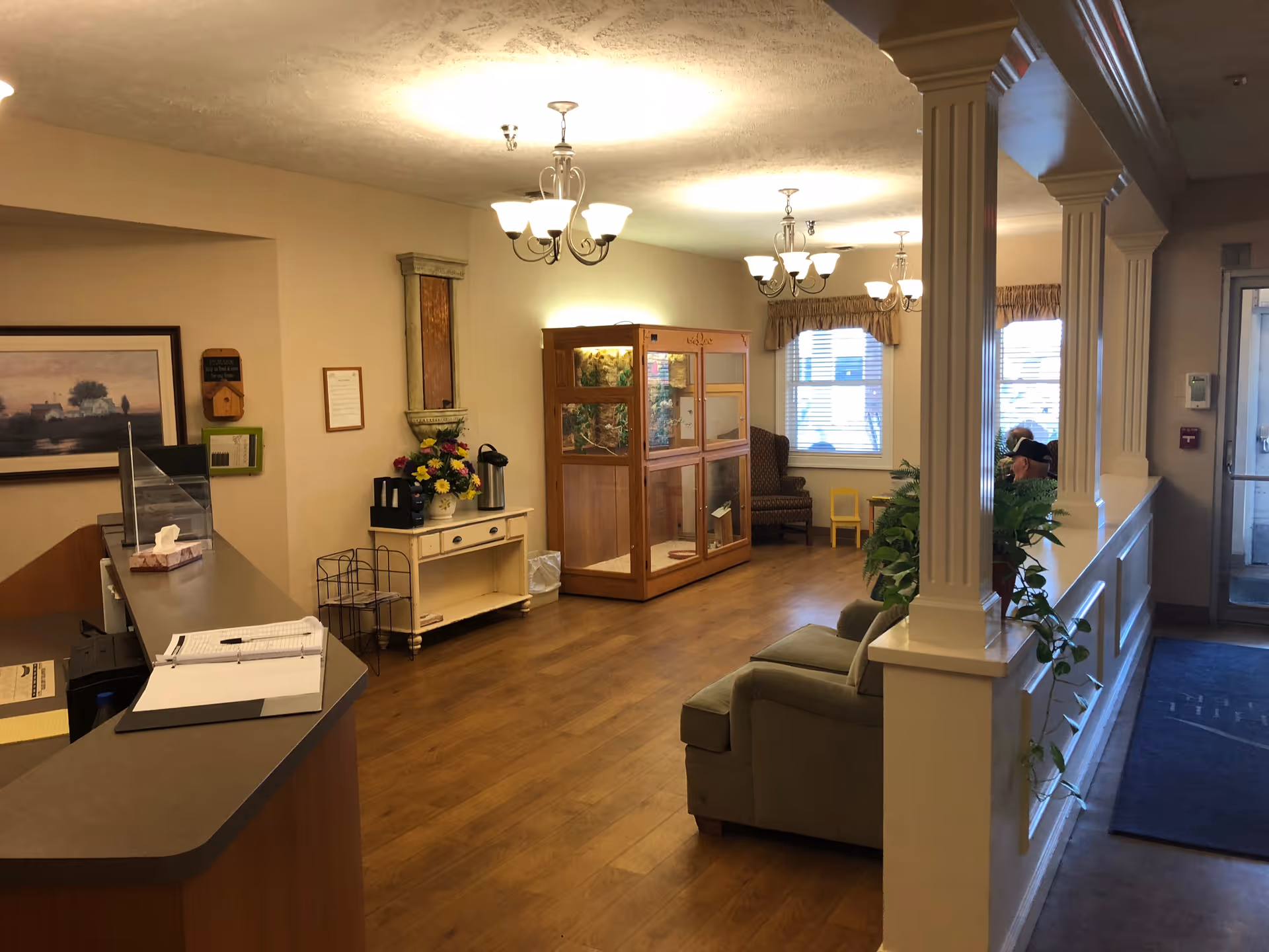 Interior view of a nursing and rehabilitation facility reception area with a front desk on the left, a small table with flowers and a coffee maker, a glass display case, seating area with armchairs, plants, and windows with blinds letting in natural light.