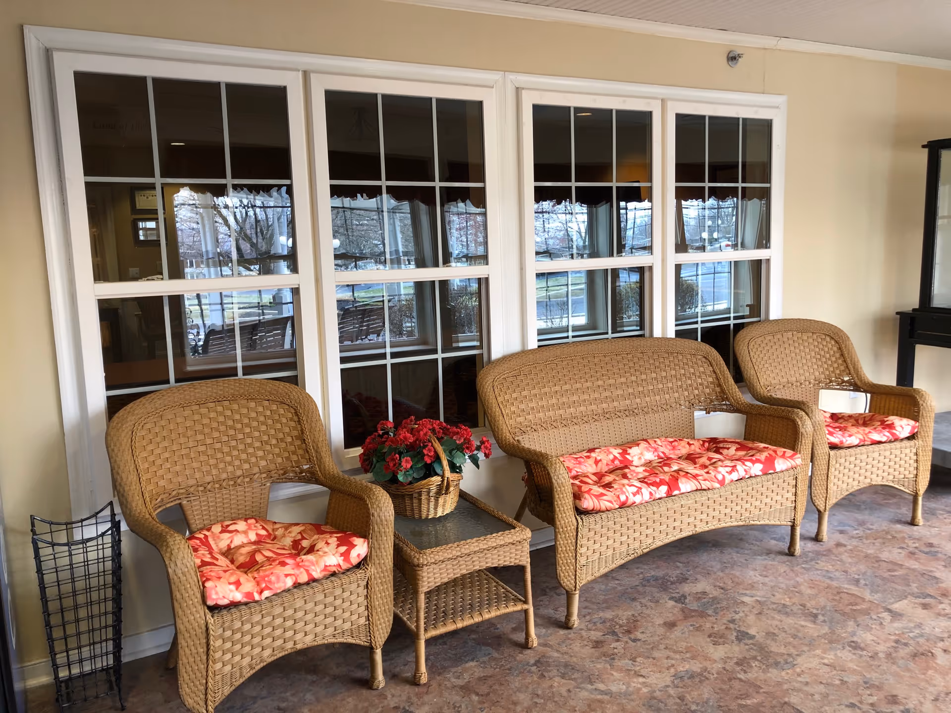 Indoor seating area with three wicker chairs and a wicker loveseat, all with red floral cushions. A small wicker table with a basket of red flowers is placed between the chairs. Large windows with white frames are behind the seating, reflecting the outdoor scene.