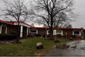 Single-story brick nursing facility with a red roof, bare trees, grassy front lawn with rocks, and a driveway on an overcast day.