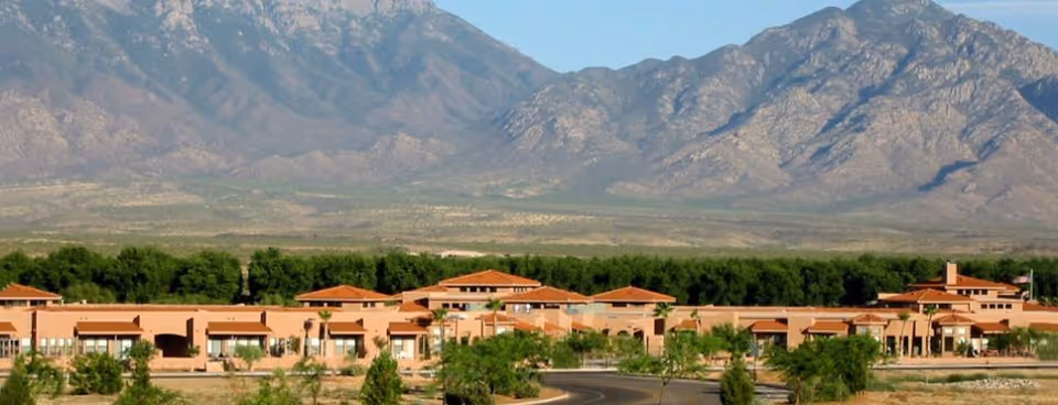 Wide exterior view of a single-story senior living facility named Silver Springs with terracotta roofs, surrounded by greenery and trees, set against a backdrop of large rocky mountains under a clear blue sky.