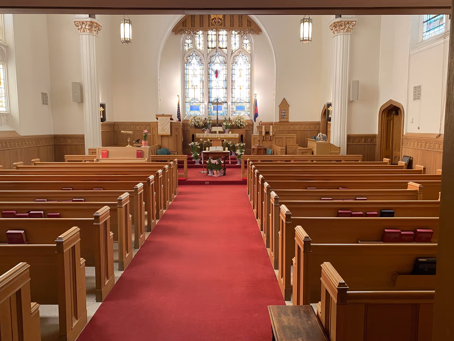 Interior view of a chapel with wooden pews arranged in rows on either side of a red carpet aisle leading to an altar adorned with flowers and religious items. The chapel features tall white columns, stained glass windows, and wooden paneling along the walls.