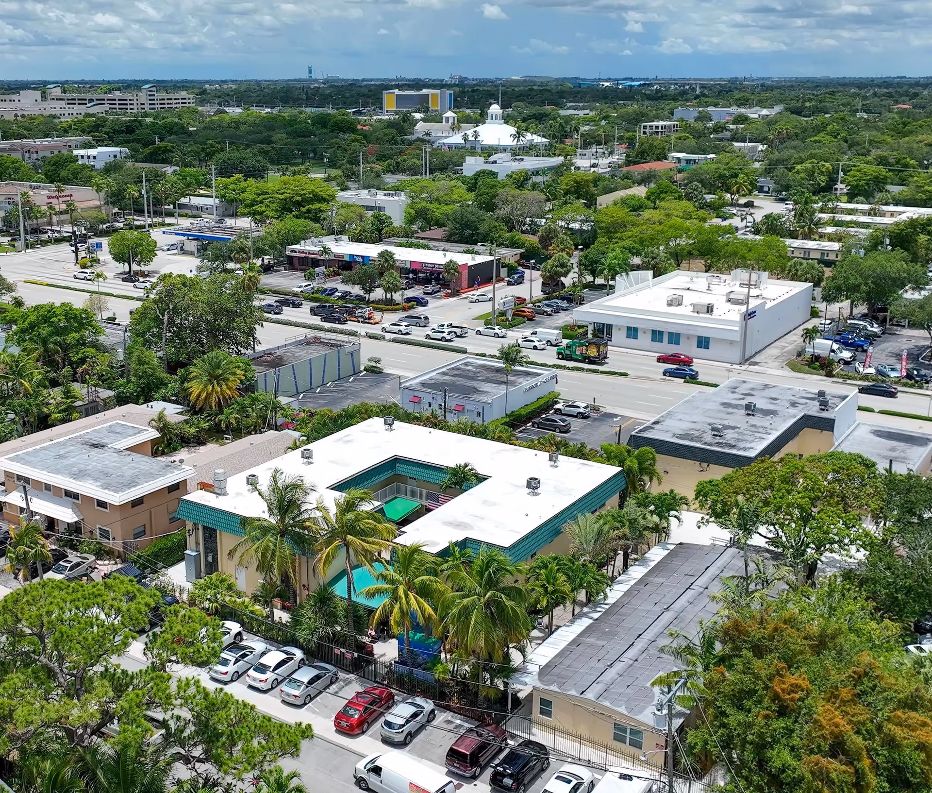 Aerial view of a neighborhood with a mix of commercial and residential buildings surrounded by trees and greenery under a partly cloudy sky. The image shows a white building with a green roof and a courtyard pool, several parked cars, and a busy street with vehicles and shops in the background.