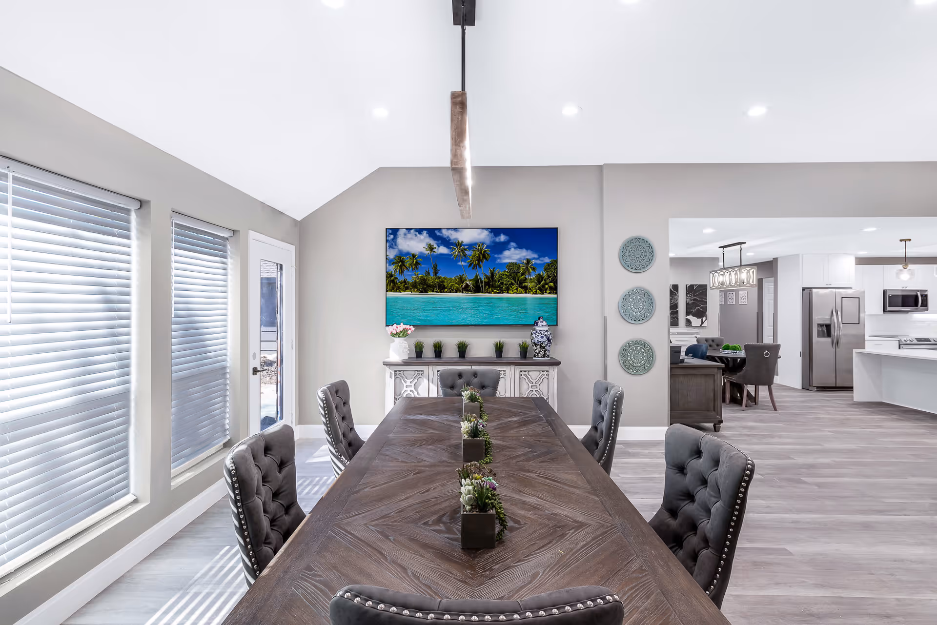 Bright open dining room with a long wooden table and upholstered chairs facing a console with a wall-mounted TV showing a tropical beach, adjacent to a modern kitchen.