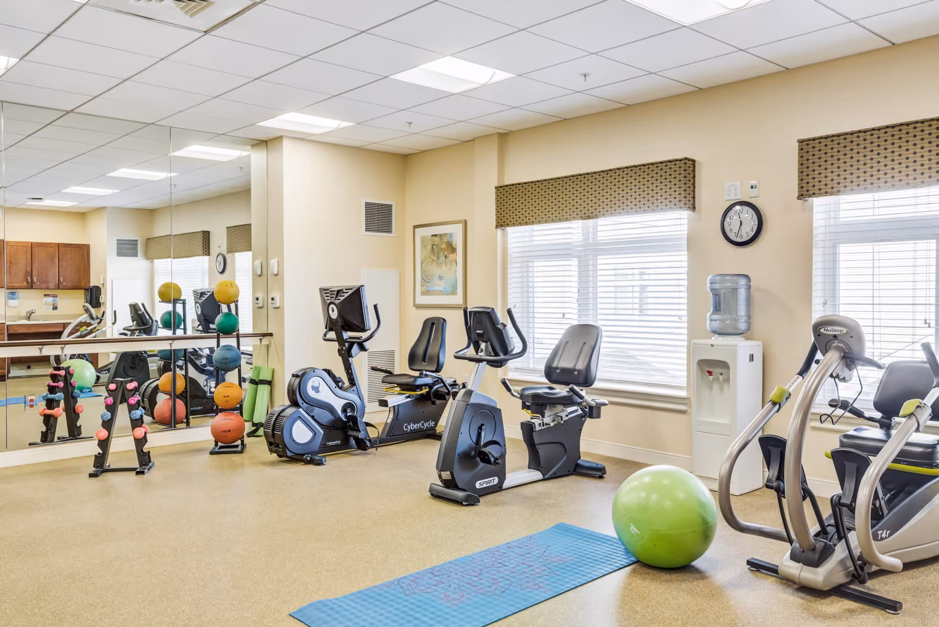 A bright fitness room in a senior living facility featuring exercise equipment including stationary bikes, an elliptical machine, a rack of colorful dumbbells, exercise balls, a yoga mat, and a water cooler. Large windows with blinds and valances allow natural light to fill the room, and a wall clock is visible above the water cooler. Mirrors cover one wall, reflecting the equipment and enhancing the sense of space.