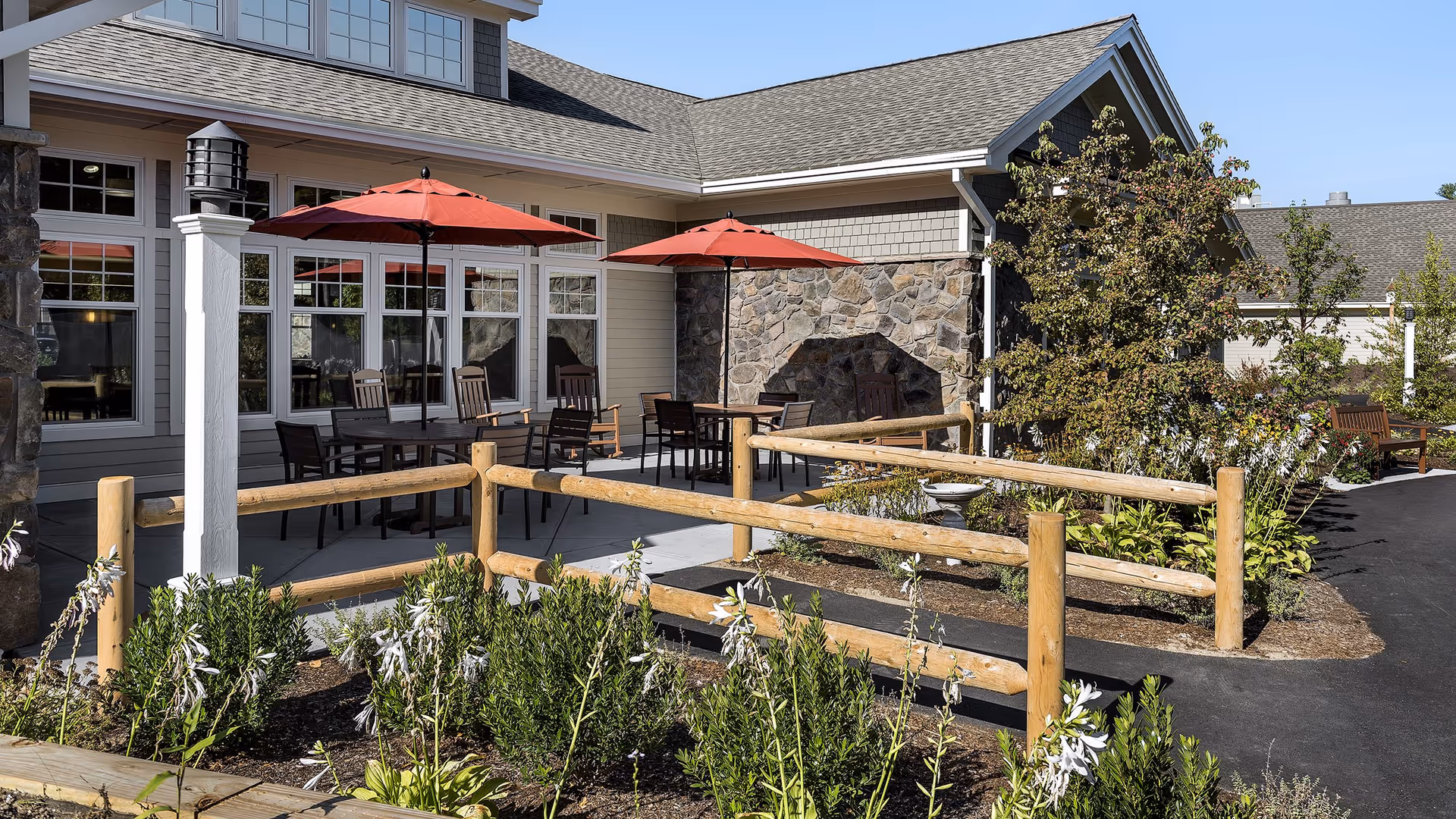 Outdoor patio area at Bridges by EPOCH at Nashua featuring tables with red umbrellas, wooden chairs, a stone wall, wooden fencing, and surrounding greenery and flowers under a clear blue sky.