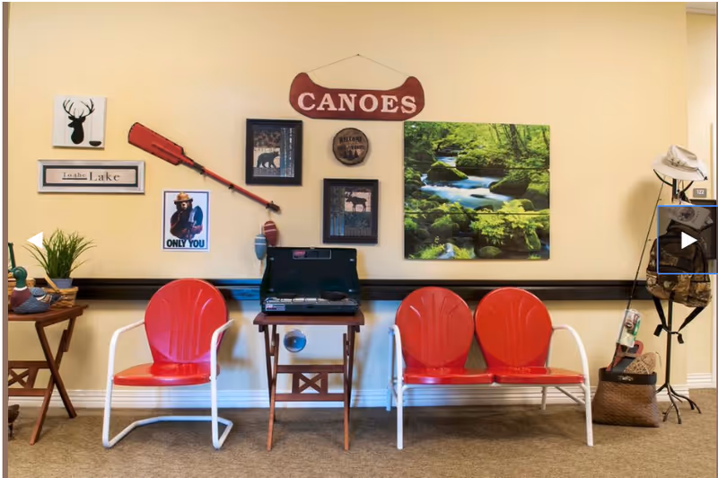 A cozy indoor seating area with two red metal chairs and a red metal bench with white frames. Between the chairs is a small wooden table holding a black portable stove. The wall behind features various decorations including a red canoe paddle, a sign that says 'CANOE', framed pictures of wildlife, a 'To the Lake' sign, and a nature landscape painting of a forest stream. To the right, there is a stand with hats and bags. To the left, a small wooden table holds a plant and decorative items.