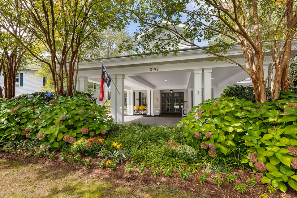 Entrance of Arbor Terrace Asheville senior living facility with a covered driveway, white pillars, green double doors, and a North Carolina state flag. The entrance is surrounded by lush green bushes, flowering plants, and trees.