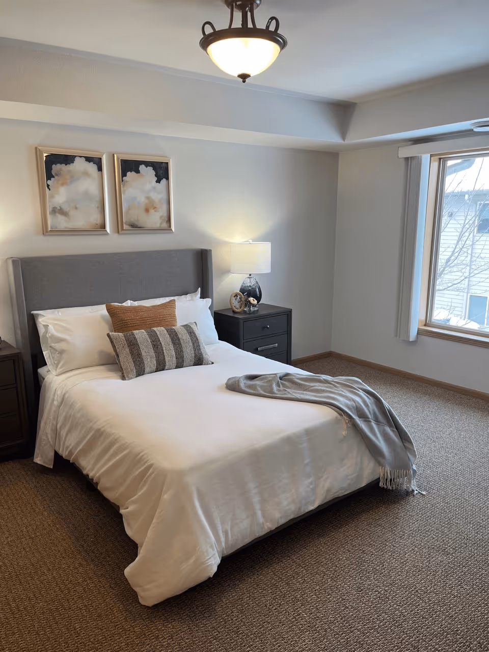 A neatly made bed with white linens, two decorative pillows, and a gray throw blanket draped on the corner. The bed has a gray upholstered headboard and is flanked by two dark wooden nightstands. One nightstand has a lamp with a white shade and a small framed photo. Above the bed are two framed abstract cloud paintings. A large window with vertical blinds lets in natural light, and the room has beige carpet and light-colored walls.
