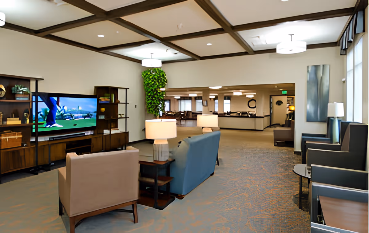 A spacious senior living facility common area with comfortable seating including armchairs and sofas facing a large flat-screen TV mounted on a wooden shelving unit. The room features a coffered ceiling with recessed lighting, a tall green plant on the wall, and additional seating along the windows with side tables and lamps. The area extends into another room with more seating and a reception desk visible in the background.